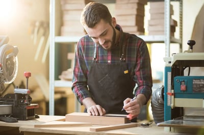 Carpintero trabajando en taller de madera, midiendo y marcando pieza sobre mesa de trabajo.