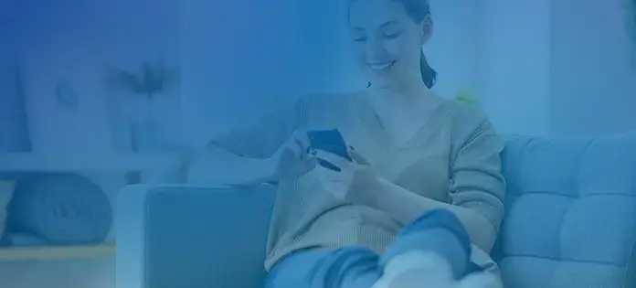 Mujer joven sonriendo mientras usa su teléfono móvil sentada en un sofá.