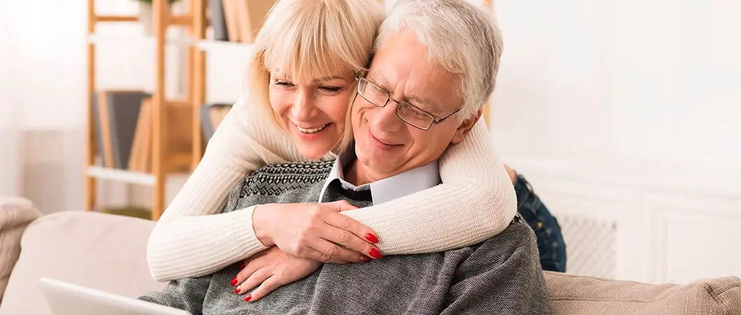Mujer abrazando por detrás a un hombre mayor sentado en un sofá, ambos sonriendo mientras miran una pantalla