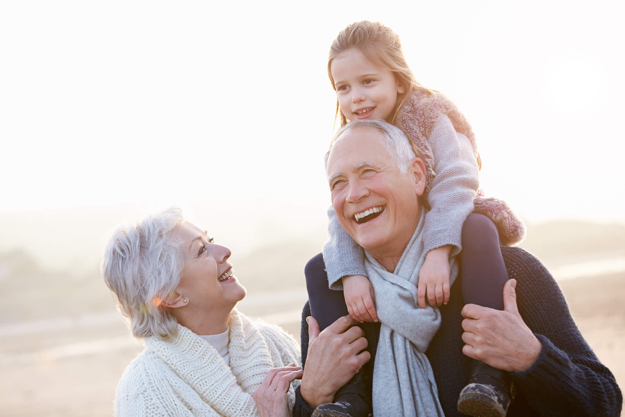 Abuelos sonrientes paseando con su nieta, quien está sentada sobre los hombros del abuelo.