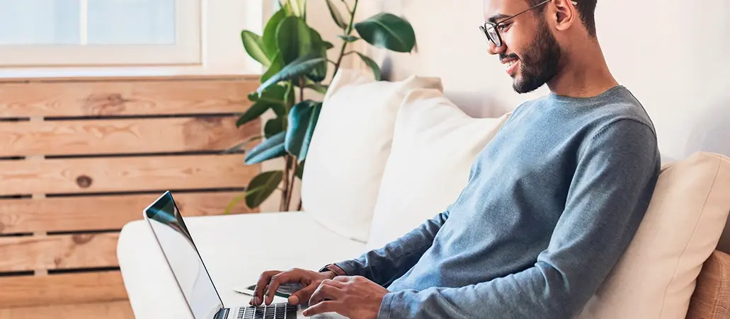 Hombre joven con gafas usando una laptop mientras sonríe sentado en un sofá