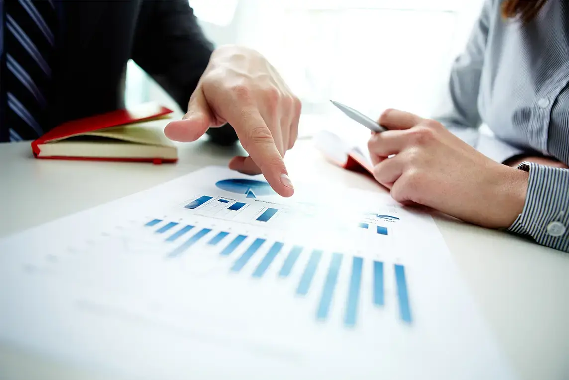 Dos personas revisando un gráfico de barras en un informe financiero sobre una mesa blanca, con una libreta roja al fondo.